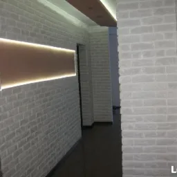 Korytarz z wykończonymi ścianami imitującymi białą cegłę, podświetlony panel dekoracyjny na ścianie, widok z perspektywy wejścia.