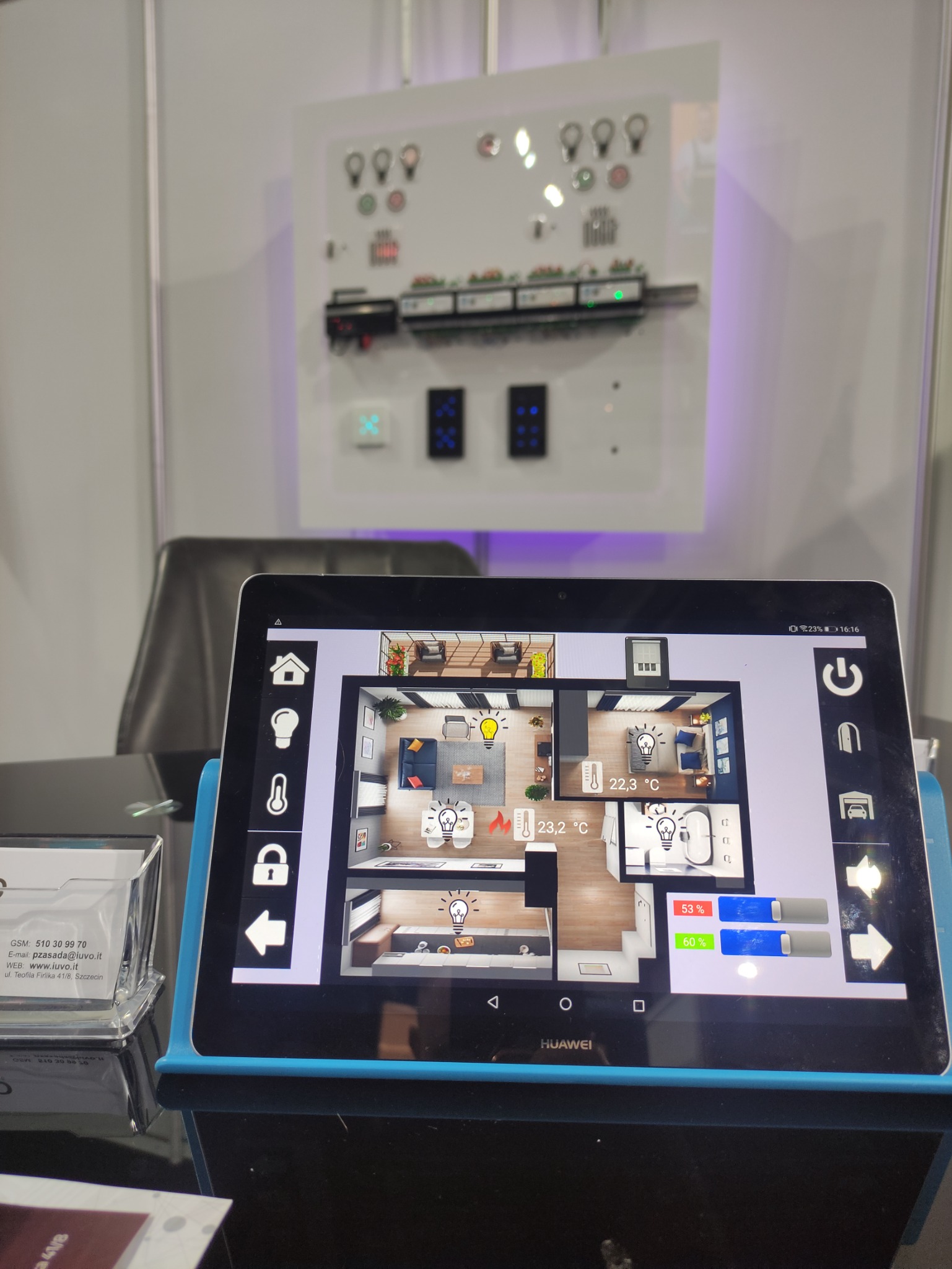 Tablet prezentujący interfejs użytkownika systemu zarządzania domem z wizualizacją planu mieszkania i kontrolą oświetlenia, na tle panelu sterowania z przyciskami i diodami LED.