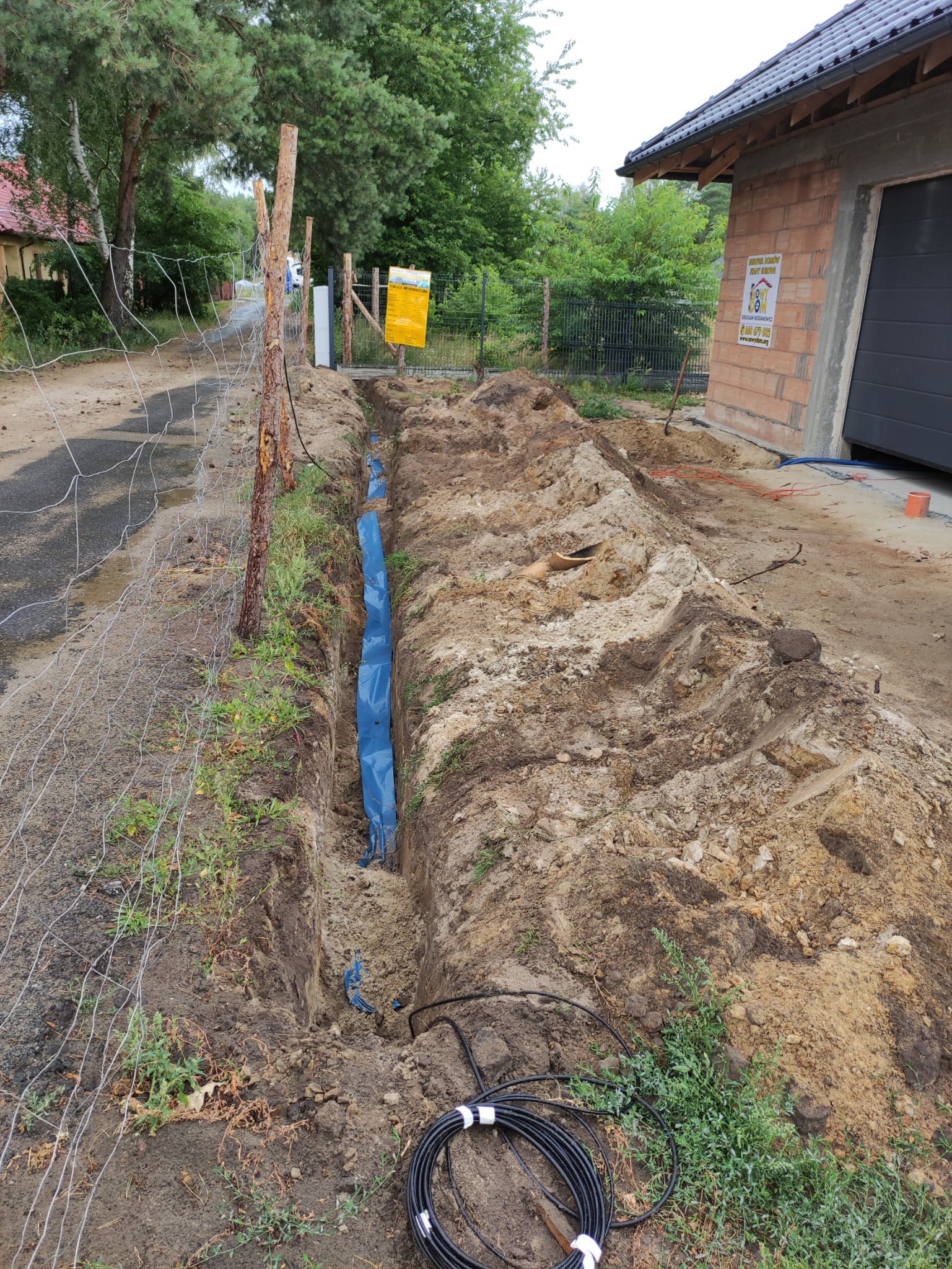 Wykop w ziemi z niebieską folią zabezpieczającą, przygotowany pod ułożenie kabli elektrycznych; na pierwszym planie zwój czarnego kabla.