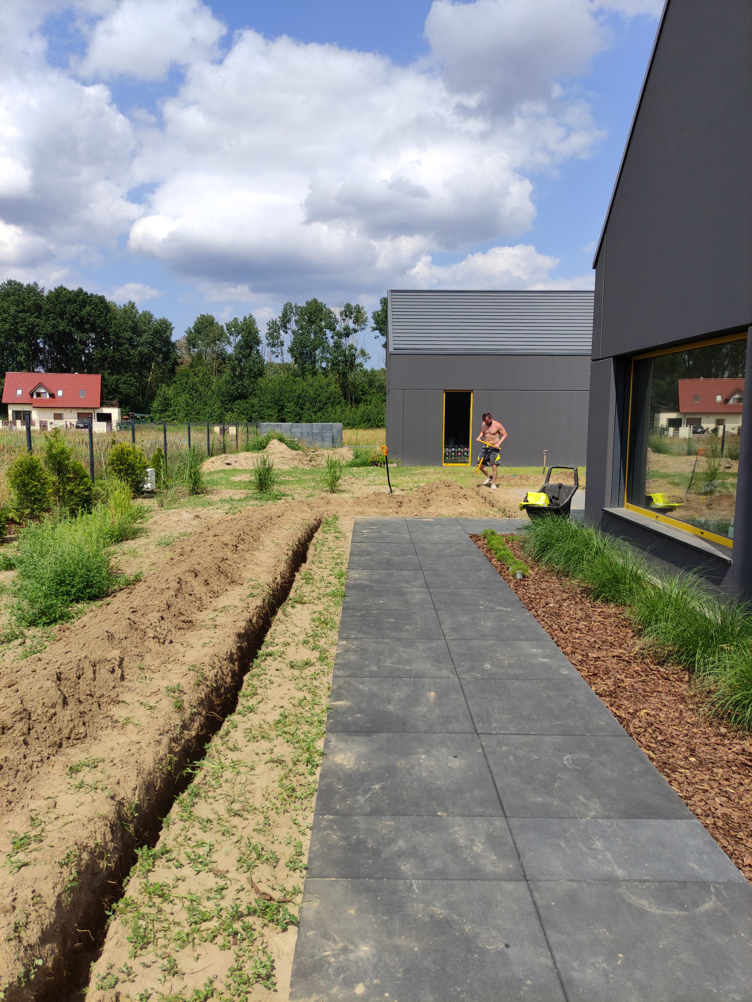 Wykopany rów w ziemi obok nowoczesnego domu z szarymi ścianami, mężczyzna kopie łopatą, widoczna kosiarka w tle, błękitne niebo z chmurami.