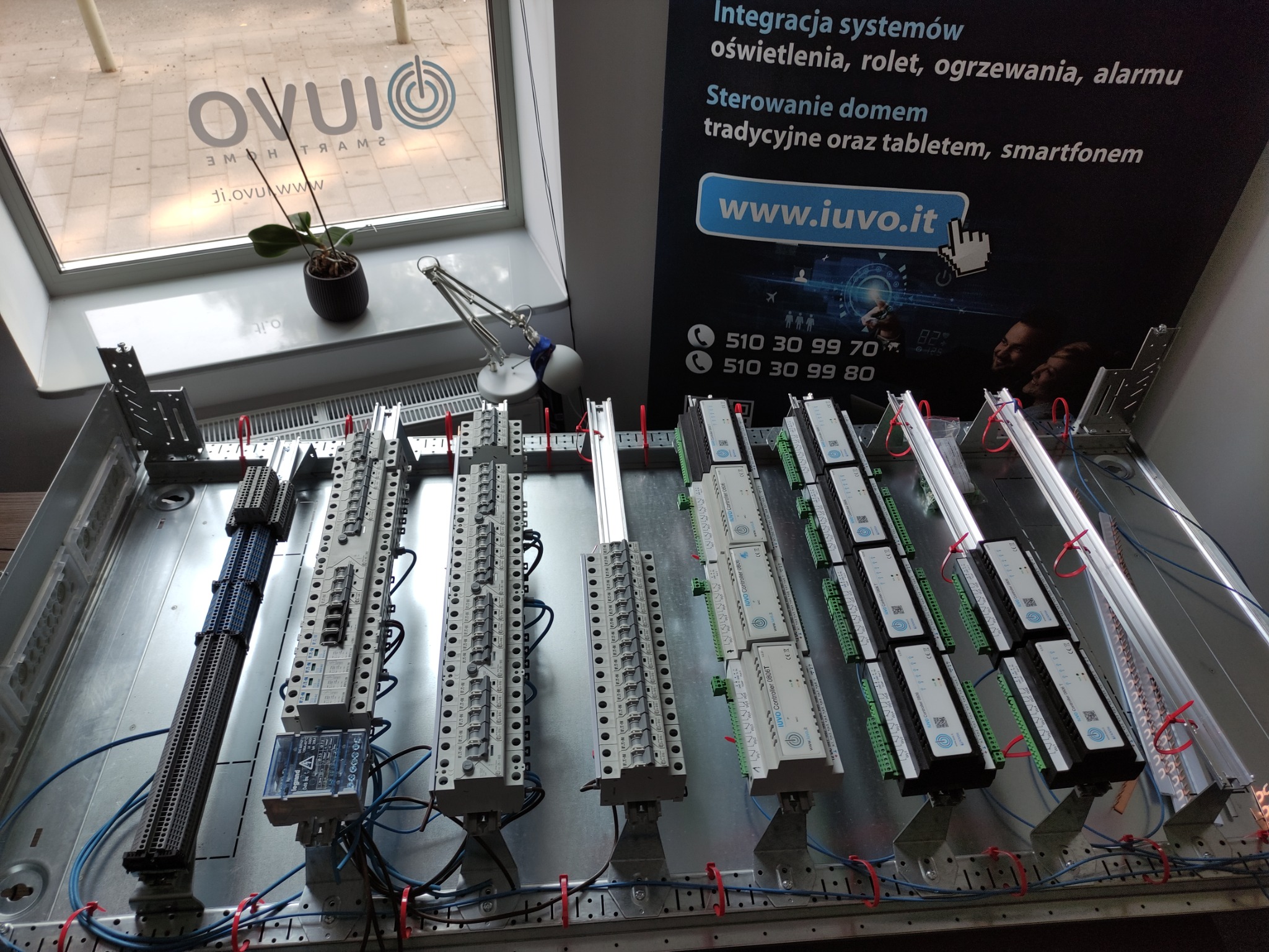 Szafa sterownicza w trakcie montażu, widoczne listwy zaciskowe, moduły sterujące i okablowanie, w tle okno z naklejonym logo firmy i plakat reklamowy z hasłem o integracji systemów.
