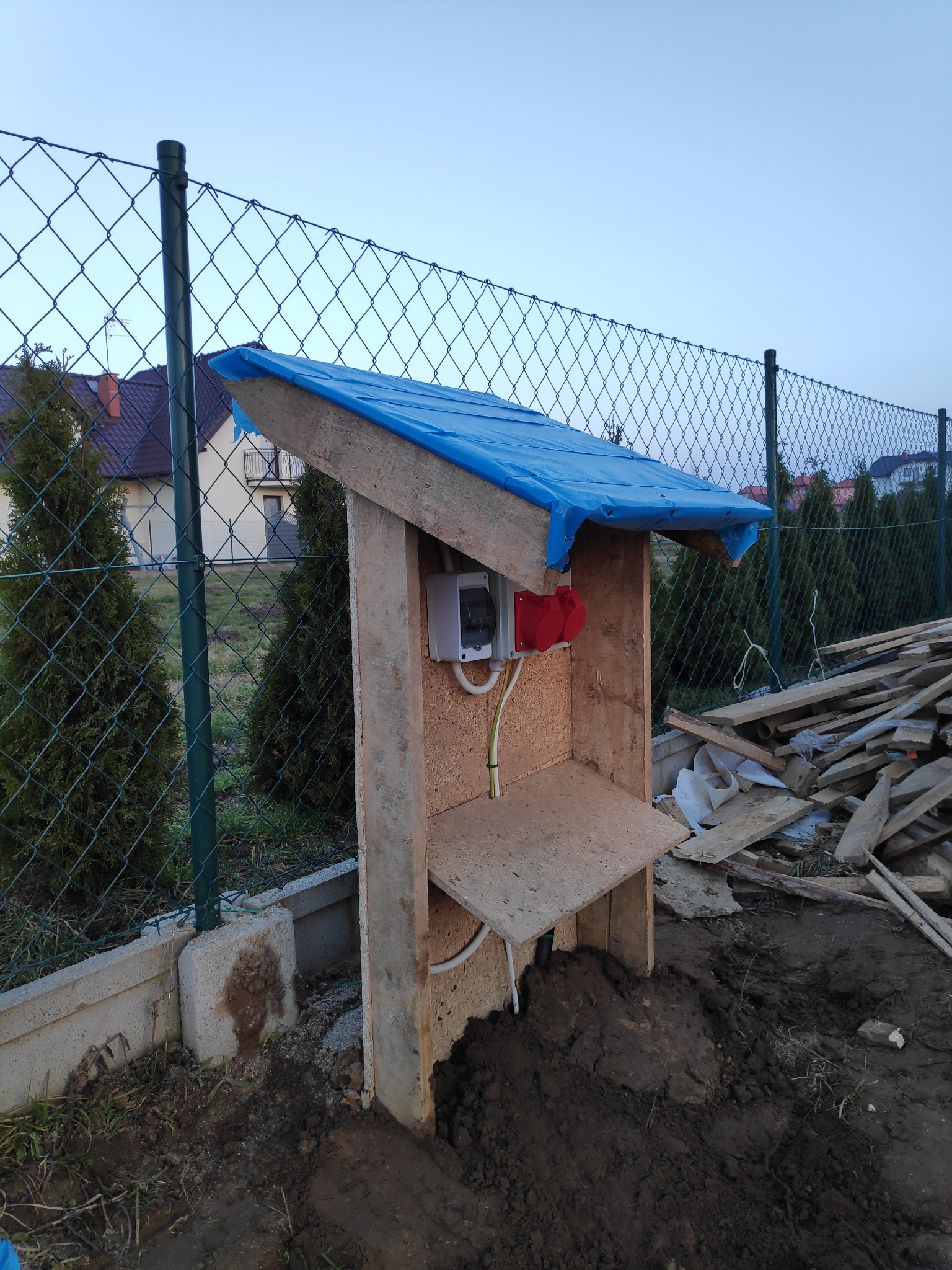 Tymczasowa skrzynka elektryczna z gniazdami siłowymi, zabezpieczona niebieską plandeką, w otoczeniu placu budowy i ogrodzenia z siatki.