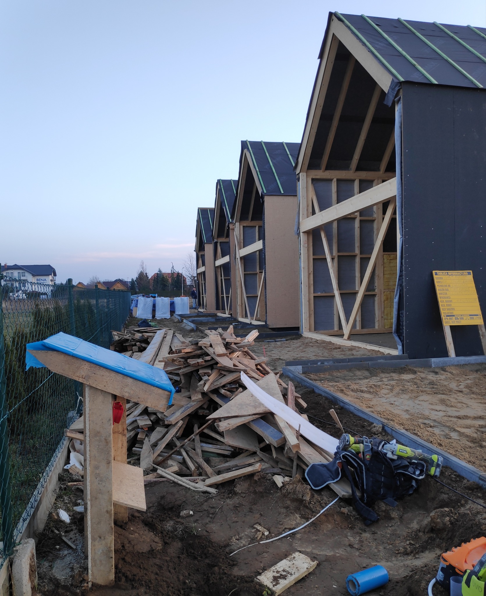 Szereg szkieletowych domków w trakcie budowy, widoczne drewniane konstrukcje, czarna membrana dachowa, tablica informacyjna budowy, oraz stos drewna i narzędzia budowlane na pierwszym planie.