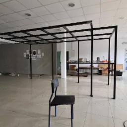 Czarna, metalowa konstrukcja przestrzenna z kratownicą na suficie, w jasnym pomieszczeniu warsztatowym z krzesłem na pierwszym planie.