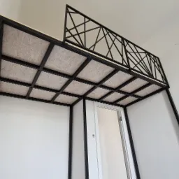 Metalowa konstrukcja antresoli z dekoracyjną balustradą, widok od dołu na sufit z płyty OSB i białe ściany.