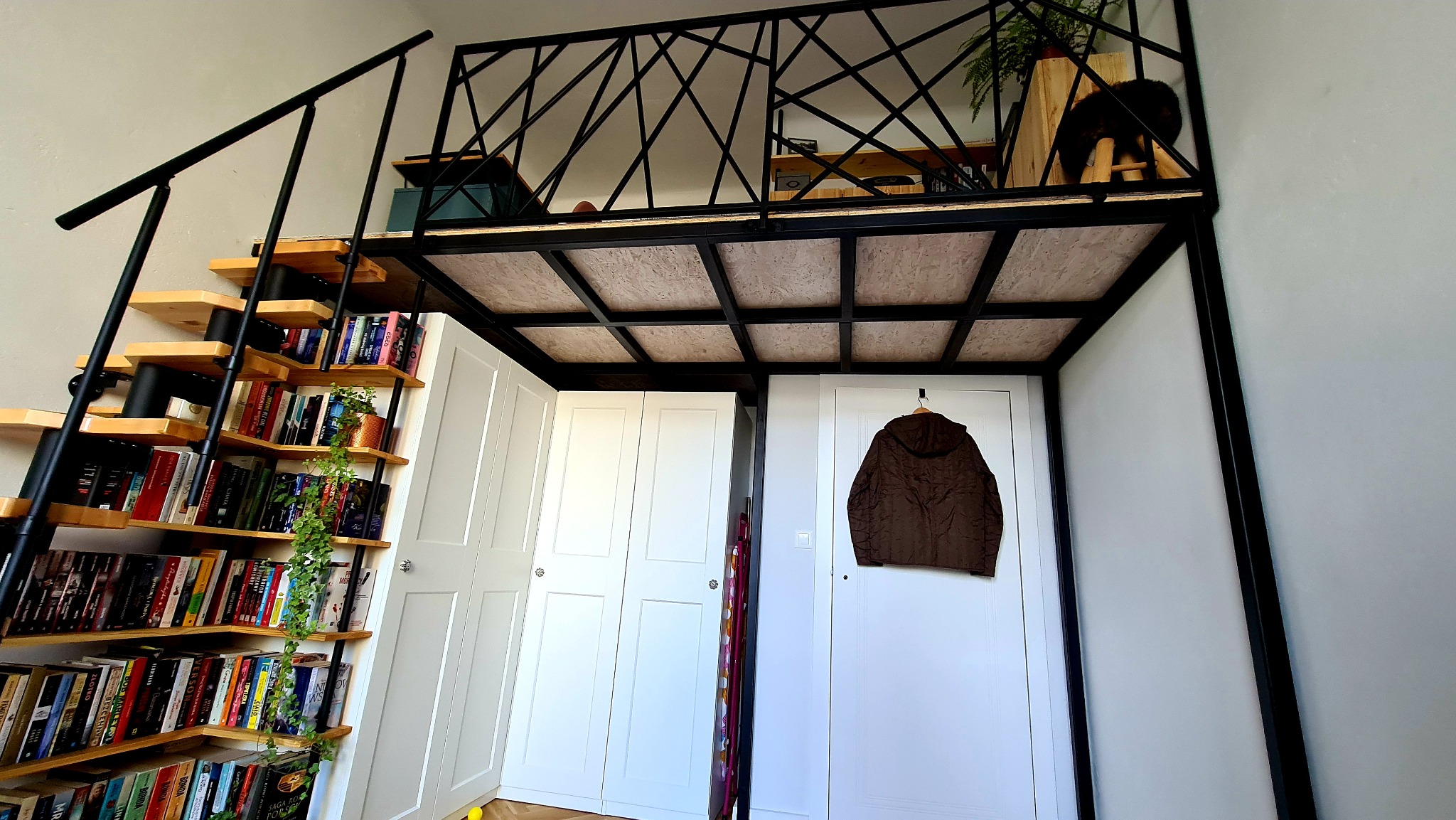 Wnętrze pokoju z antresolą na metalowej konstrukcji, drewnianymi schodami z półkami na książki i białymi szafami wnękowymi pod spodem. Na antresoli widoczne rośliny i przedmioty dekoracyjne.