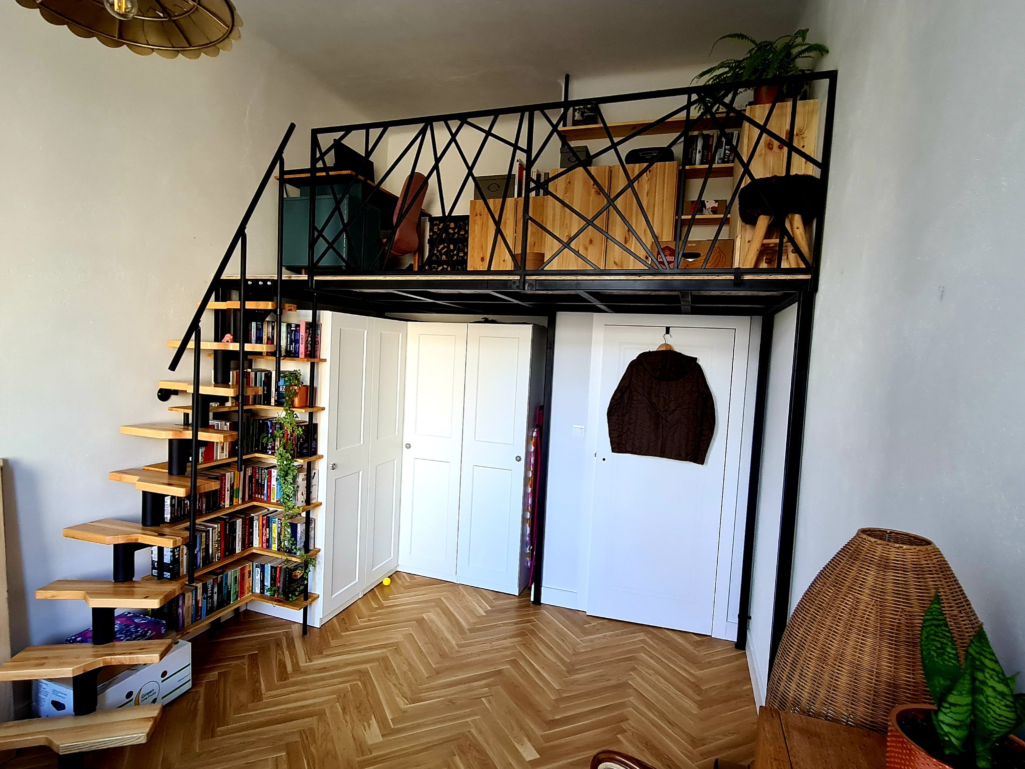 Wnętrze mieszkania z antresolą na konstrukcji metalowej, drewniane schody z półkami na książki, białe szafy wnękowe pod antresolą, parkiet w jodełkę.