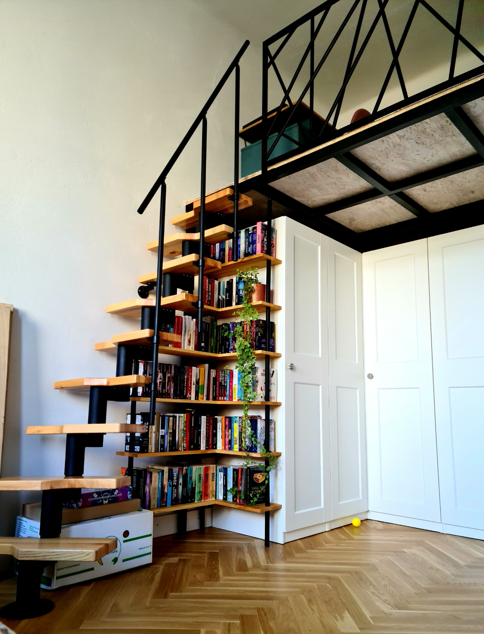 Ażurowe schody z drewnianymi stopniami i czarną metalową konstrukcją, pełniące funkcję regału na książki, prowadzące na antresolę z metalową konstrukcją i białymi szafami w tle, całość na parkiecie...