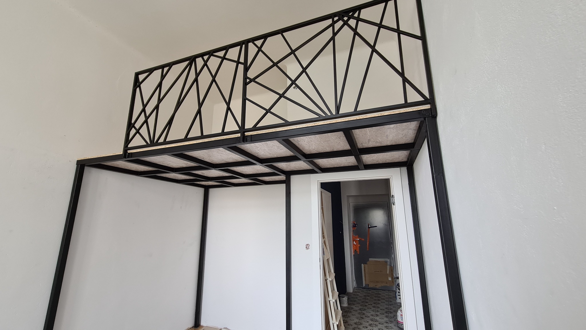 Metalowa konstrukcja antresoli z balustradą o geometrycznym wzorze, widoczna w surowym wnętrzu z białą ścianą i otwartymi drzwiami do kolejnego pomieszczenia.