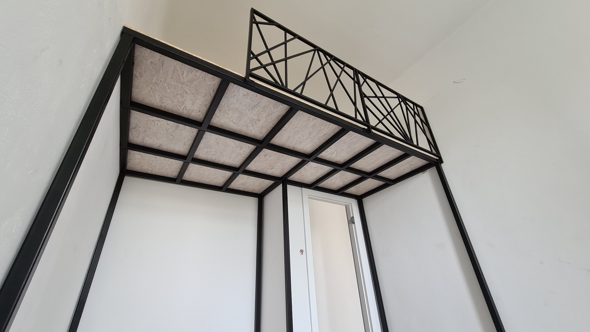 Metalowa konstrukcja antresoli z dekoracyjną balustradą, widok od dołu na sufit z płyty OSB i białe ściany.