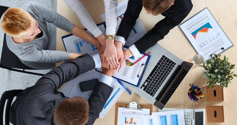 Zespół biznesowy łączy dłonie nad stołem z laptopem, dokumentami z wykresami i długopisami, symbolizując współpracę i jedność w biurze.