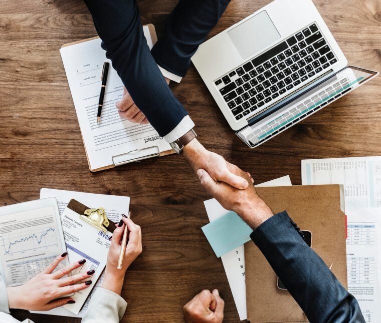 Uścisk dłoni nad drewnianym biurkiem z laptopem, dokumentami i tabletem, symbolizujący zawarcie umowy lub porozumienia biznesowego.