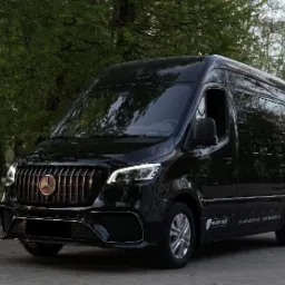 Czarny, luksusowy bus marki Mercedes z chromowanym grillem i logo, zaparkowany na tle zieleni miejskiej.
