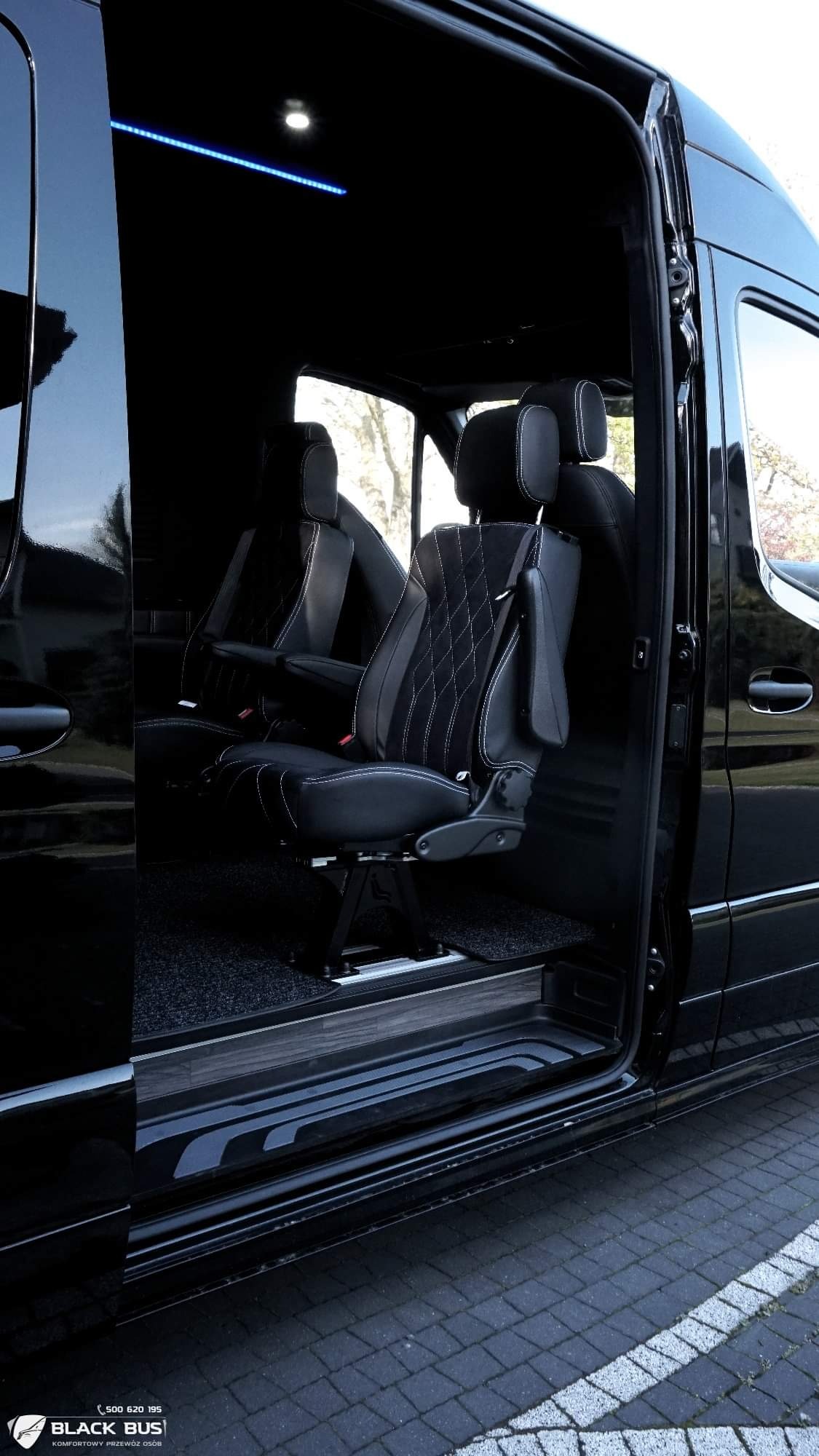 Wnętrze czarnego busa z otwartymi drzwiami przesuwnymi, ukazujące skórzane fotele z białymi przeszyciami i regulowanymi podłokietnikami, podświetlenie LED na suficie.