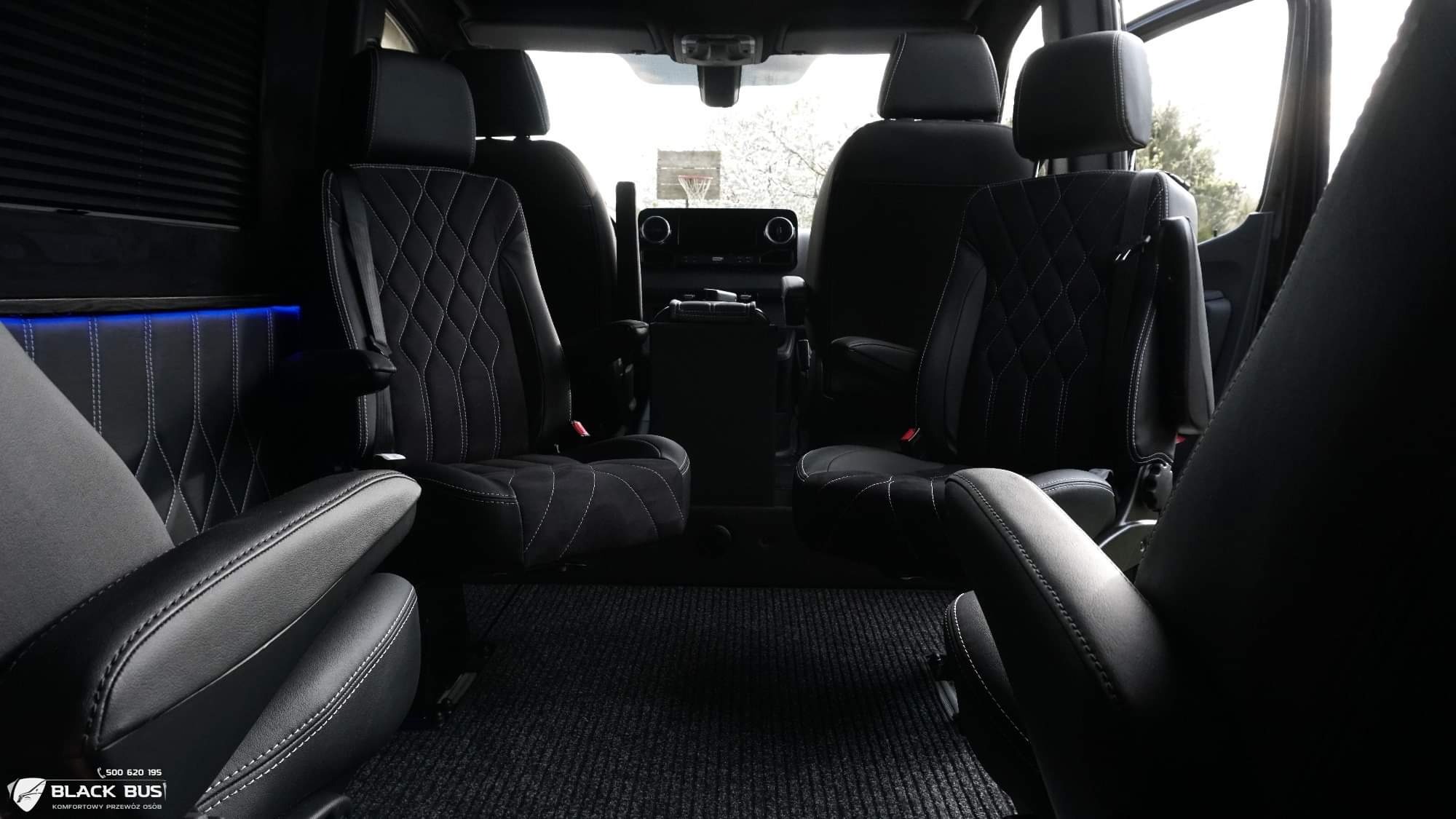 Wnętrze luksusowego busa z czarną skórzaną tapicerką, pikowanymi fotelami z białymi przeszyciami i subtelnym niebieskim oświetleniem LED.