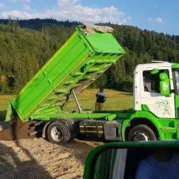 Zielona wywrotka Scania z otwartą skrzynią ładunkową wysypuje ziemię na nierówny teren; w tle zalesione wzgórza i postać mężczyzny.