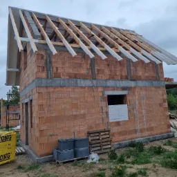 Budowa domu jednorodzinnego: ściany z czerwonej cegły, częściowo wykonana konstrukcja dachu z drewnianymi belkami, widoczne materiały budowlane na paletach.