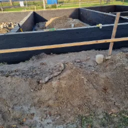 Zabezpieczone fundamenty budynku, pomalowane na czarno, z widoczną izolacją przeciwwilgociową i drewnianym szalunkiem, ziemia wokół wykopu.