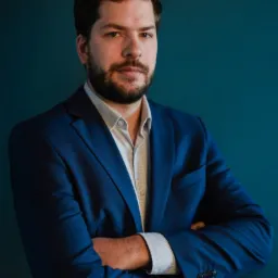 Portret mężczyzny z brodą w niebieskim garniturze, stojącego na tle jednolitego, ciemnoturkusowego tła z założonymi rękami.