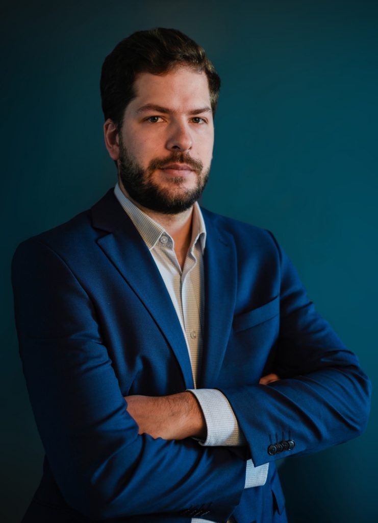 Portret mężczyzny z brodą w niebieskim garniturze, stojącego na tle jednolitego, ciemnoturkusowego tła z założonymi rękami.