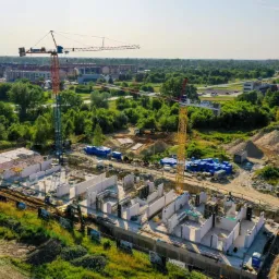 Wysoka perspektywa na plac budowy z dwoma żurawiami wieżowymi, widoczne fundamenty i pierwsze ściany z betonu komórkowego, przygotowane do dalszych prac konstrukcyjnych.