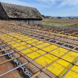 Siatka zbrojeniowa ułożona na żółtych płytach szalunkowych, z dystansami z tworzywa sztucznego, w tle drewniany budynek gospodarczy i pole uprawne.