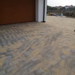 Nowo ułożona, szara kostka brukowa z piaskiem wypełniającym szczeliny przed nowoczesnym domem z brązową bramą garażową.