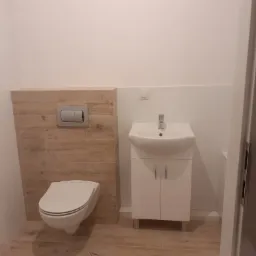 Nowoczesna łazienka z podwieszaną toaletą, umywalką z szafką i płytkami imitującymi drewno na podłodze i fragmencie ściany, widok zza uchylonych drzwi.