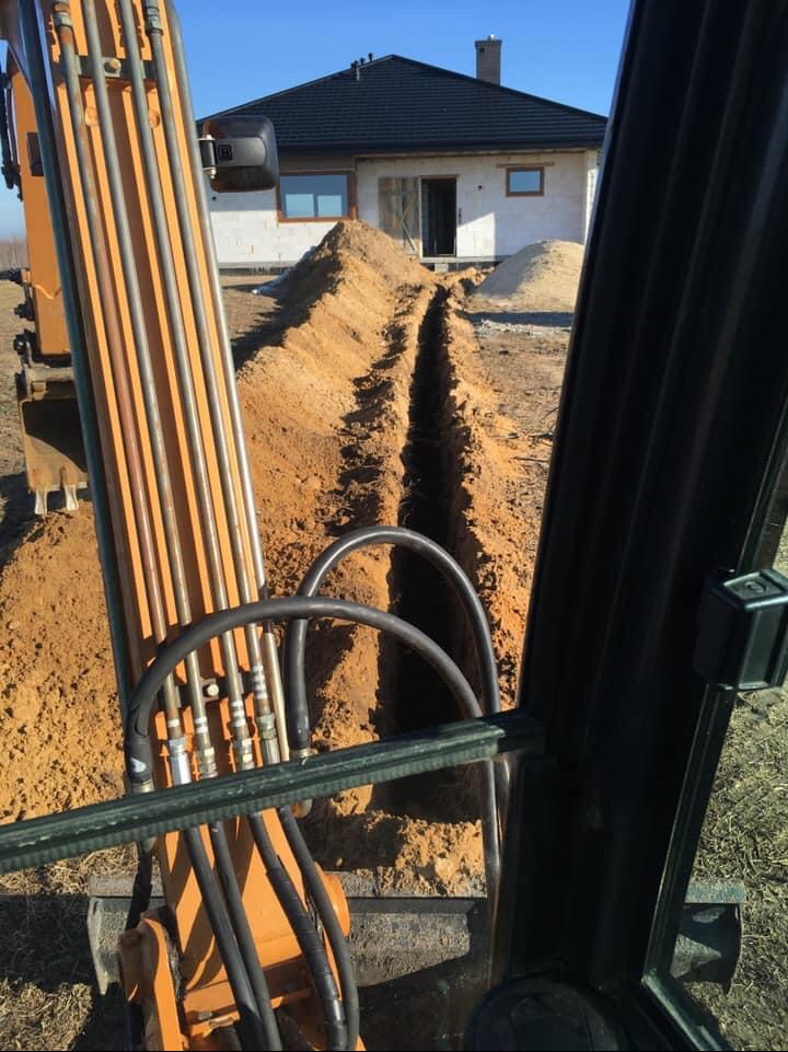 Wąski wykop w ziemi, widok z kabiny koparki, w tle budynek w stanie surowym.