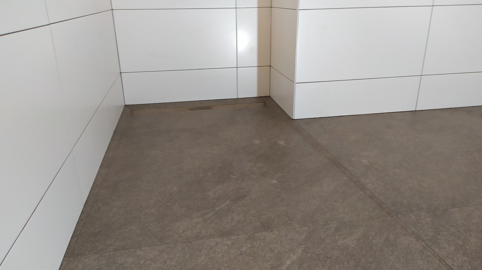 Szare płytki podłogowe w łazience z odpływem liniowym i białymi płytkami ściennymi ułożonymi poziomo.