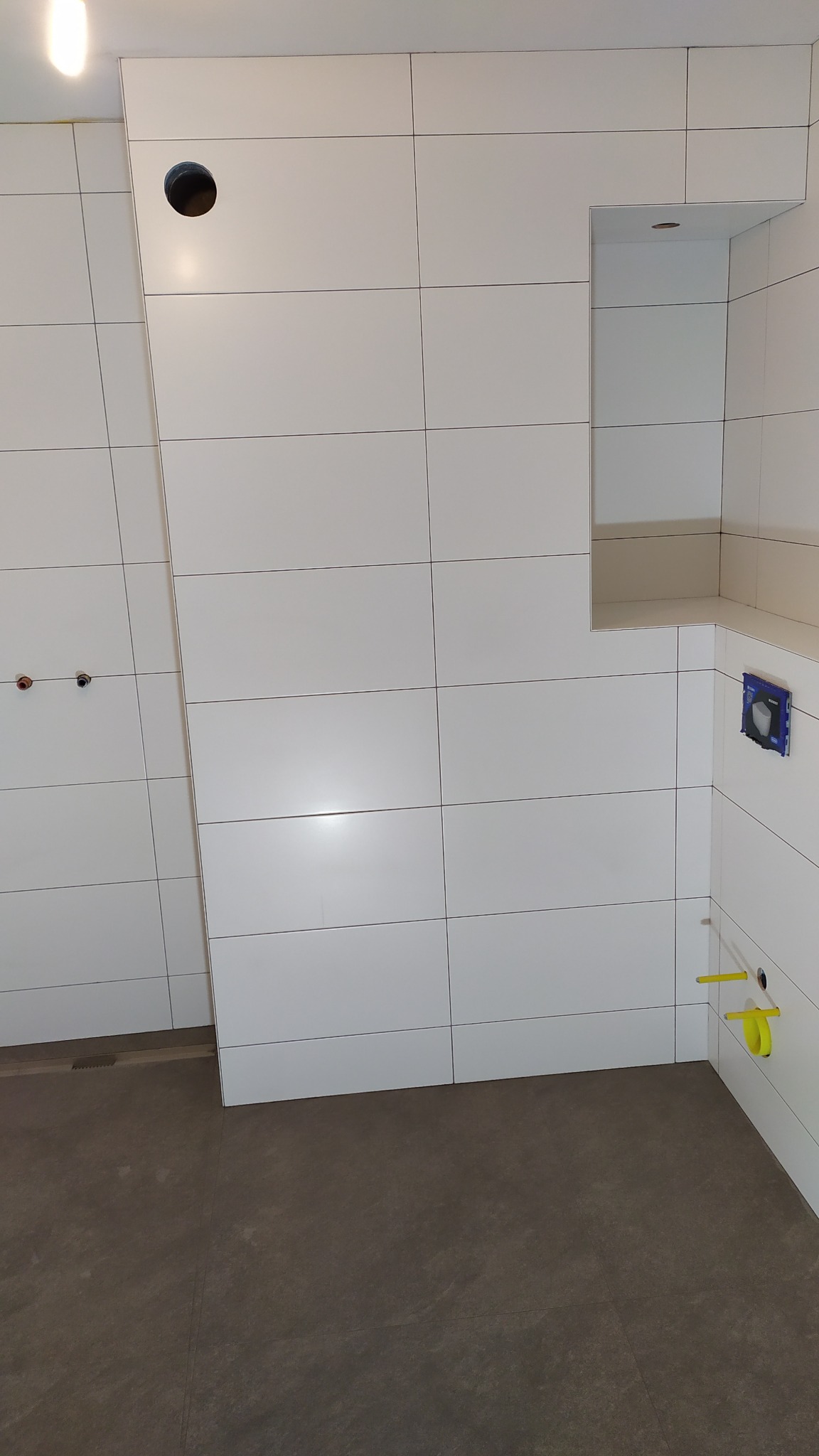 Wnętrze łazienki w trakcie prac wykończeniowych, widoczne białe, prostokątne płytki na ścianach z ciemną fugą, wnęka w ścianie z oświetleniem, przygotowane instalacje wodne i odpływ liniowy...