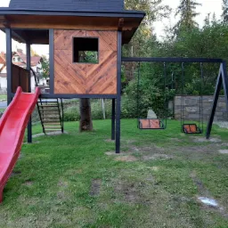 Drewniany domek na palach z czerwoną zjeżdżalnią i huśtawkami na tle zielonego trawnika i drzew