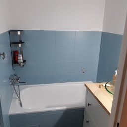 Emt Serwis Tomasz Sobutka - Niebieska łazienka z białą wanną, szklaną umywalką na blacie z jasnego drewna i chromowaną baterią. Na ścianie wiszą półki z kosmetykami.