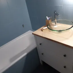 Nowoczesna łazienka z wanną, umywalką ze szklaną misą i białymi płytkami w odcieniach błękitu, uzupełniona drewnianym blatem i białymi szafkami z szufladami.