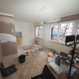 Emt Serwis Tomasz Sobutka - Pokój w trakcie prac przygotowawczych do malowania: ściany częściowo pokryte gruntem i gładzią, widoczne narzędzia, wiadro i worki z materiałami budowlanymi, podłoga zabezpieczona folią,...