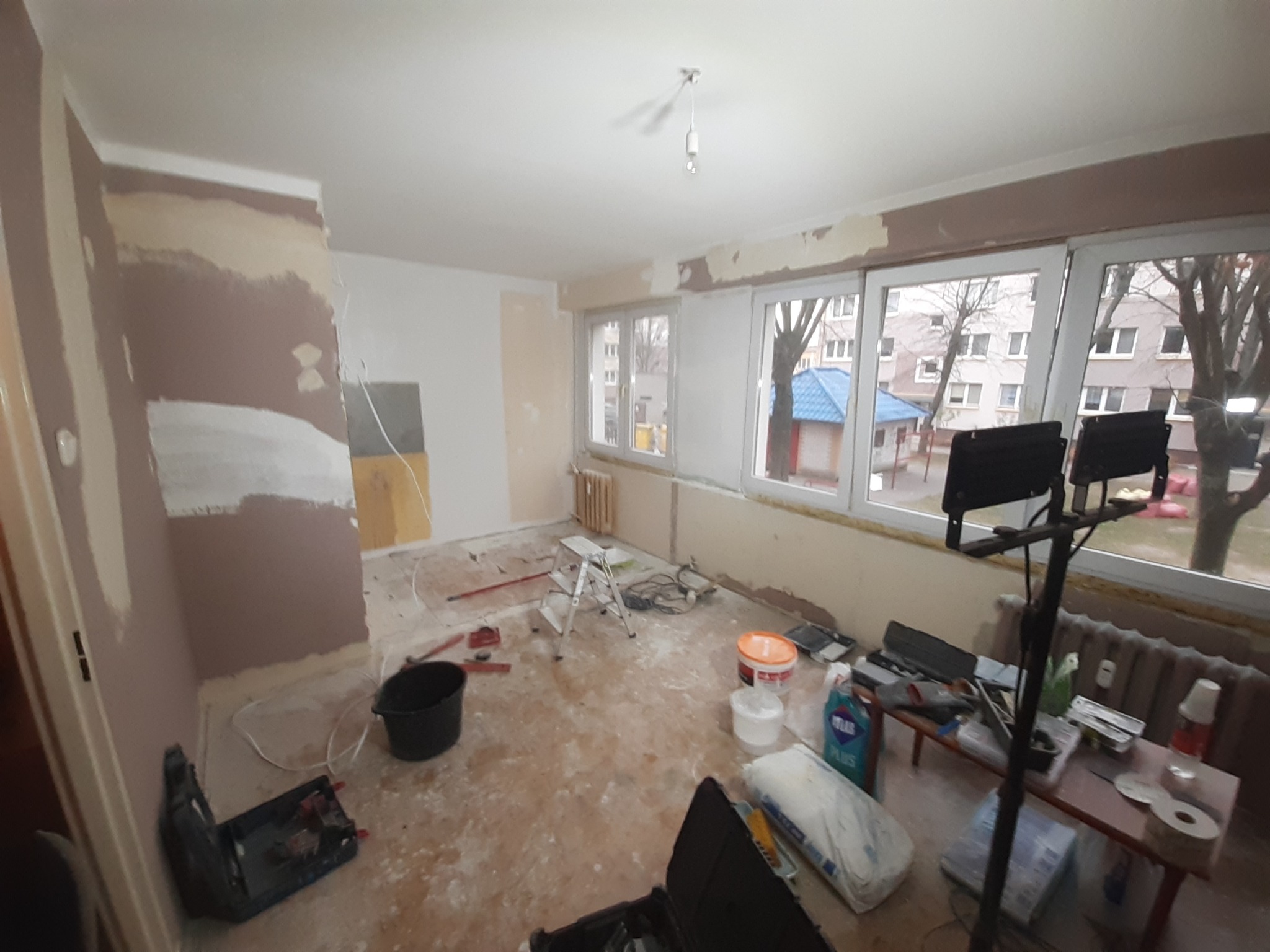 Pokój w trakcie prac przygotowawczych do malowania: ściany częściowo pokryte gruntem i gładzią, widoczne narzędzia, wiadro i worki z materiałami budowlanymi, podłoga zabezpieczona folią,...