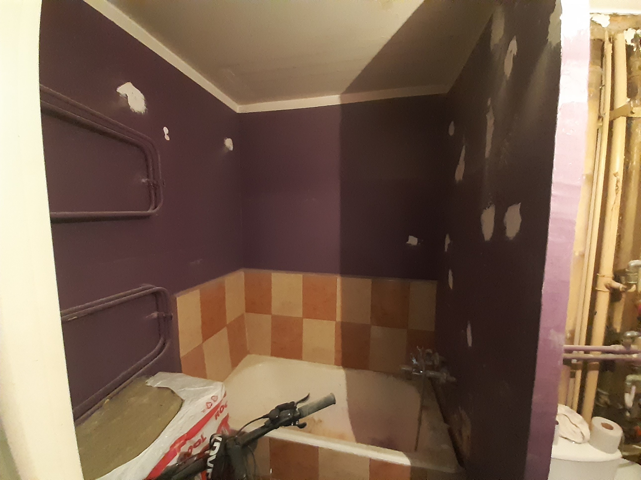 Łazienka w trakcie prac, ściany pomalowane na fioletowo z widocznymi łatami gładzi, stara wanna z kafelkami w odcieniach beżu i brązu, rury na ścianie oraz paczka wełny mineralnej przy grzejniku.