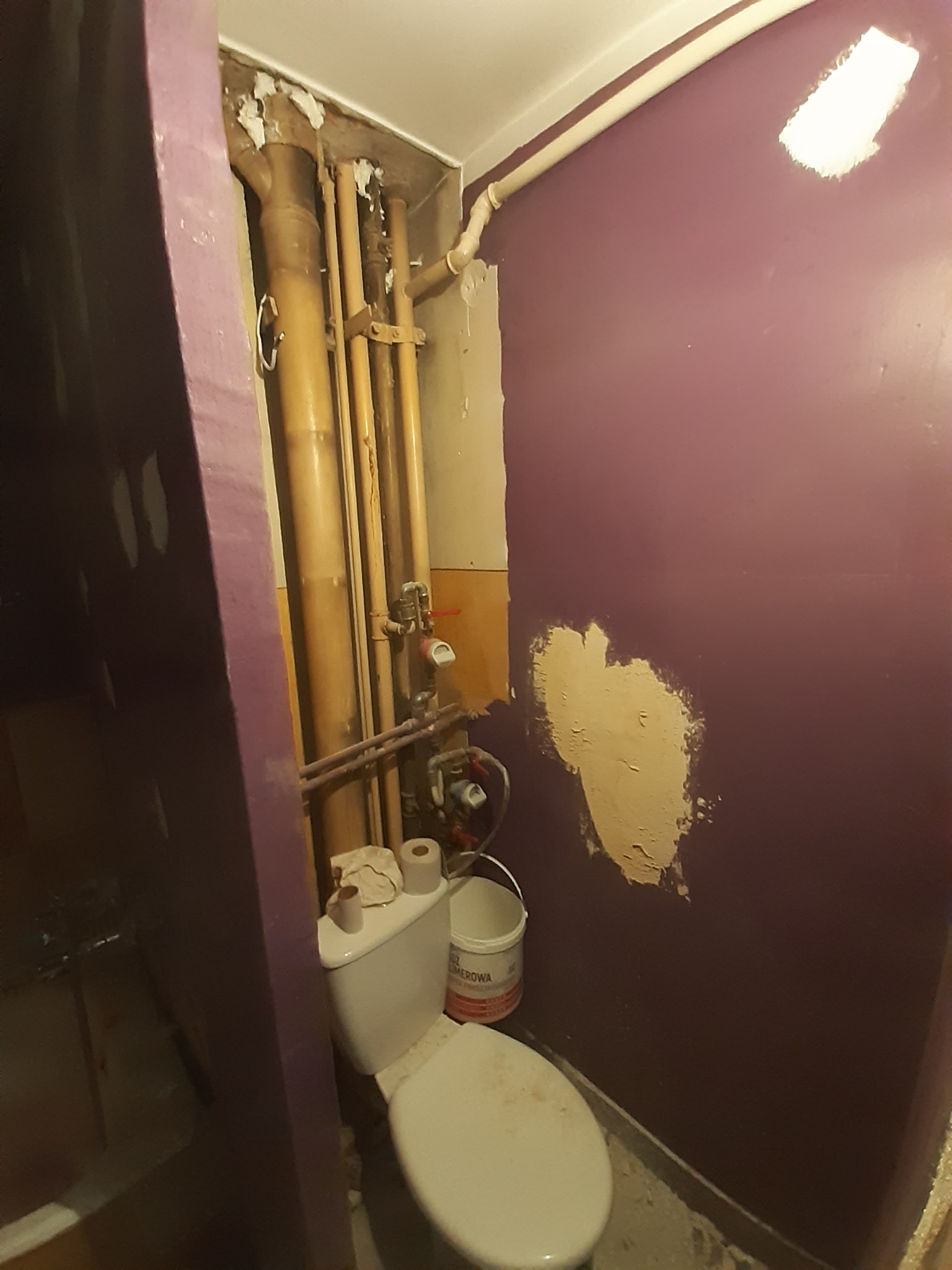 Fioletowa ściana łazienki z widocznymi śladami po usuniętych płytkach i przygotowaniem do gładzi, obok toaleta i rury centralnego ogrzewania.