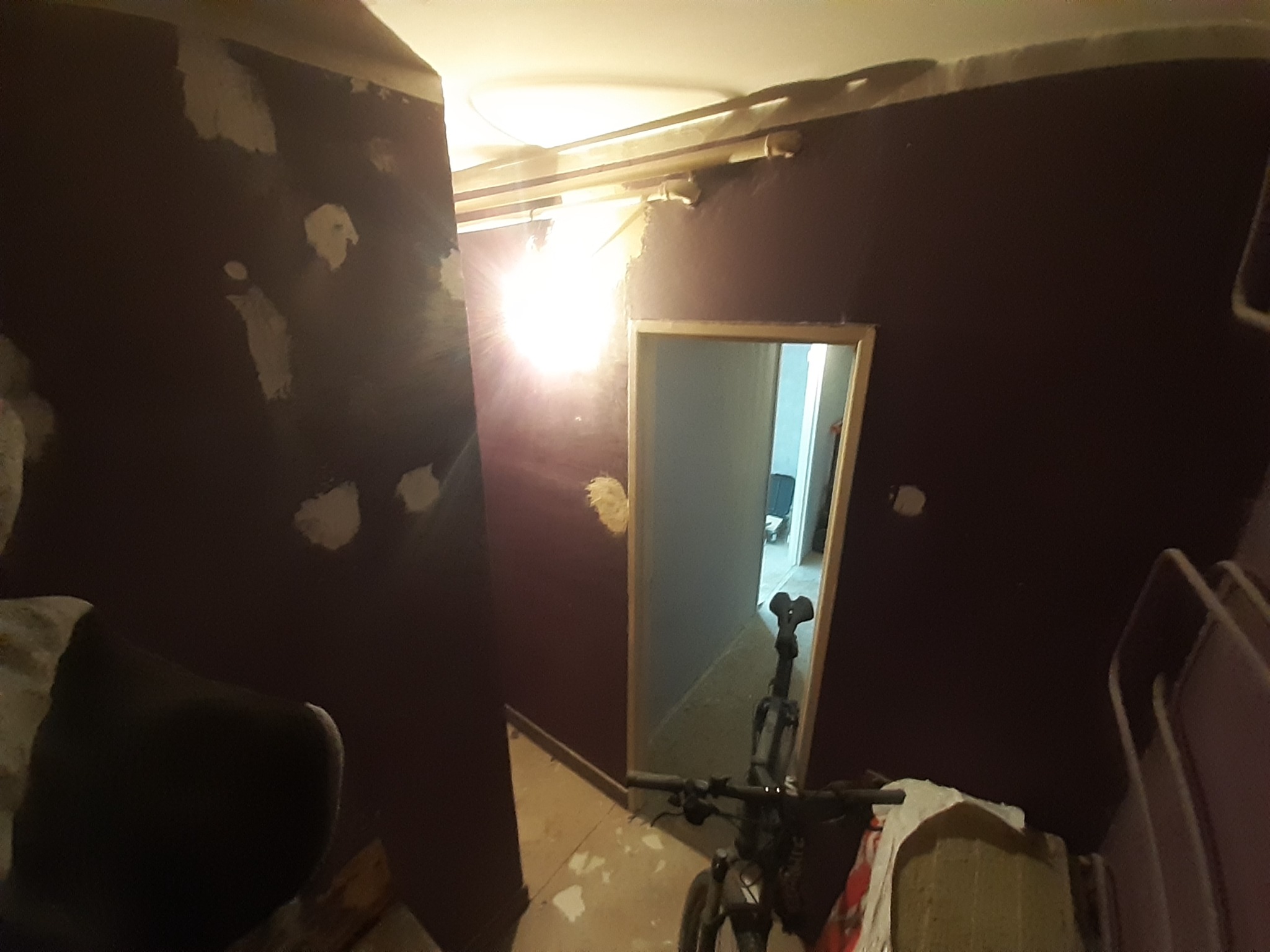 Pokój w trakcie remontu: ciemna ściana z widocznymi łatami gładzi, surowy sufit z wystającymi elementami, widok na kolejne pomieszczenie przez otwarte drzwi, rower w pomieszczeniu.