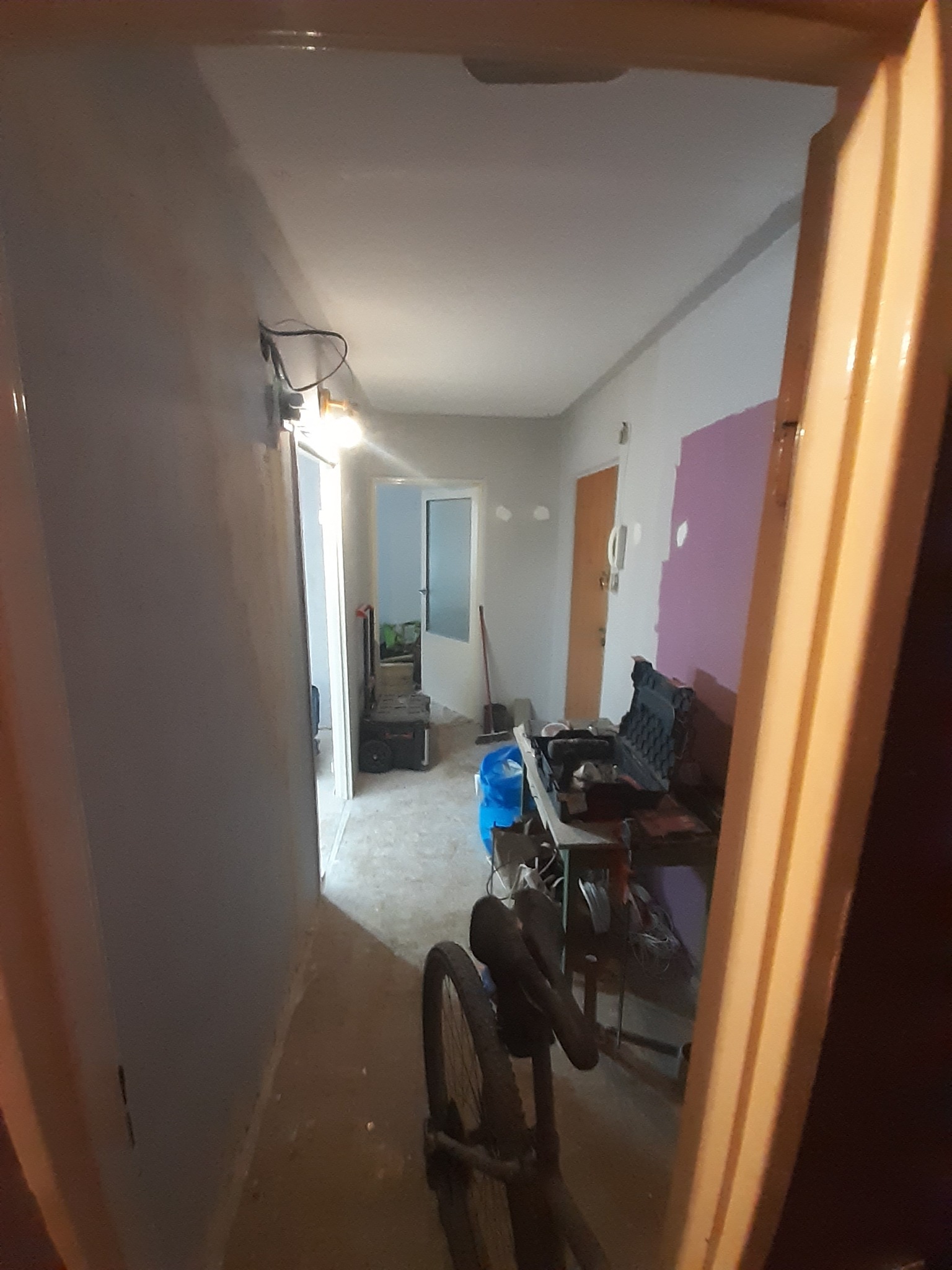Korytarz w trakcie malowania: ściany częściowo pokryte szarą farbą, widoczny fragment fioletowej ściany, narzędzia malarskie, rower w kadrze.