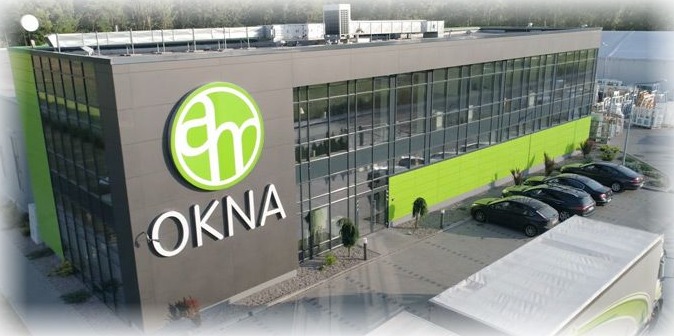 Budynek firmy AM Okna z dużym logo na fasadzie, widoczne zaparkowane samochody osobowe.