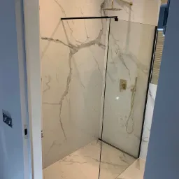 Kabina prysznicowa typu walk-in z czarnymi profilami, wykończona płytkami imitującymi marmur, z złotą armaturą, widok zza uchylonych drzwi.