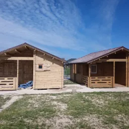 Dwa drewniane domki letniskowe z brązowymi dachami, jeden z niebieską plandeką na werandzie, na trawiastym terenie z ścieżką, pod błękitnym niebem.