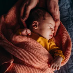 Noworodek śpi otulony koralowym kocykiem na szarym tle, dłonie rodzic&oacute;w delikatnie otaczają dziecko, widoczny zegarek na męskim nadgarstku i złota bransoletka na damskim.