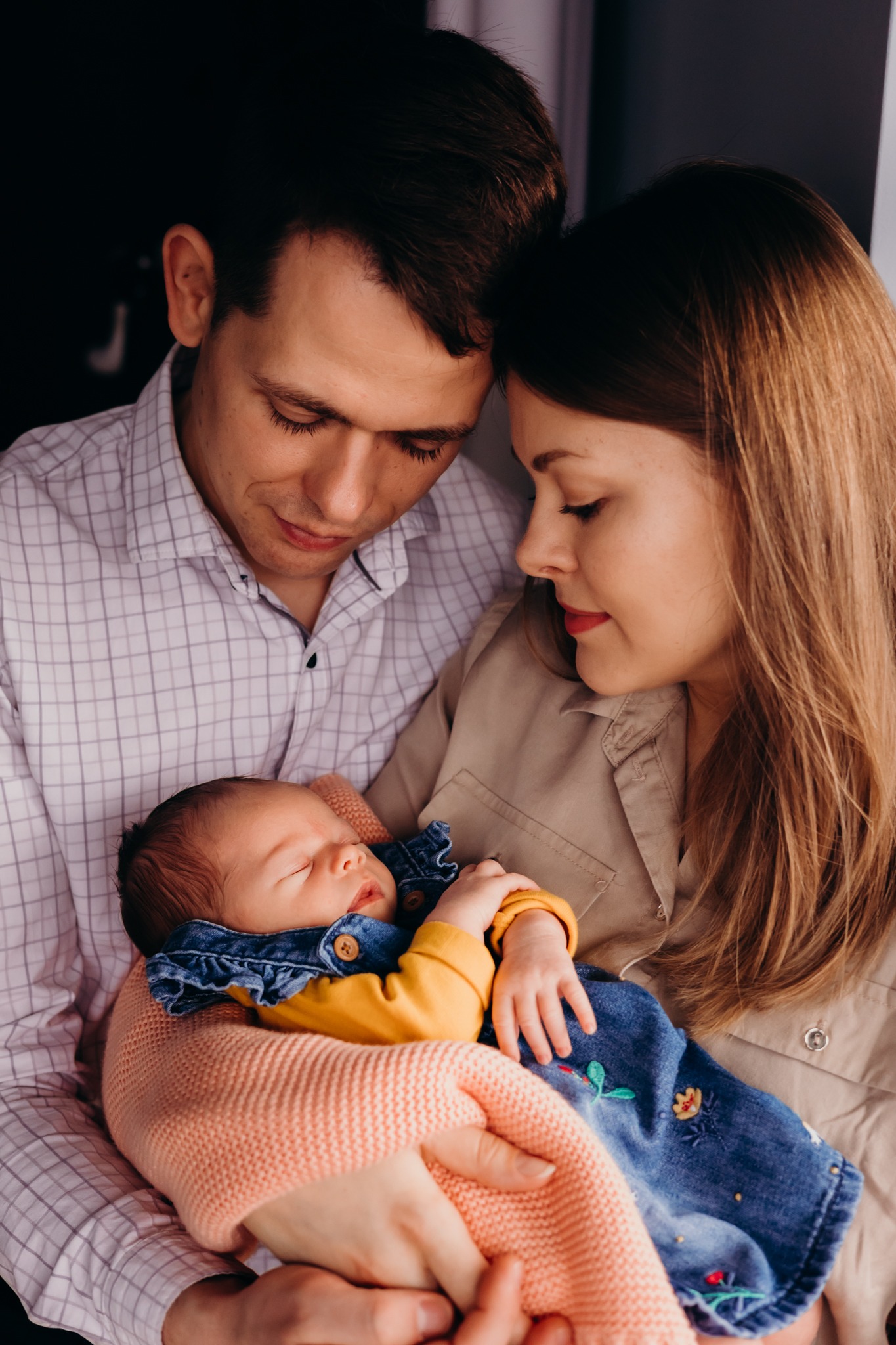 Rodzice tulą śpiącego noworodka w denimowej sukience z haftem, delikatne oświetlenie podkreśla bliskość i miłość.