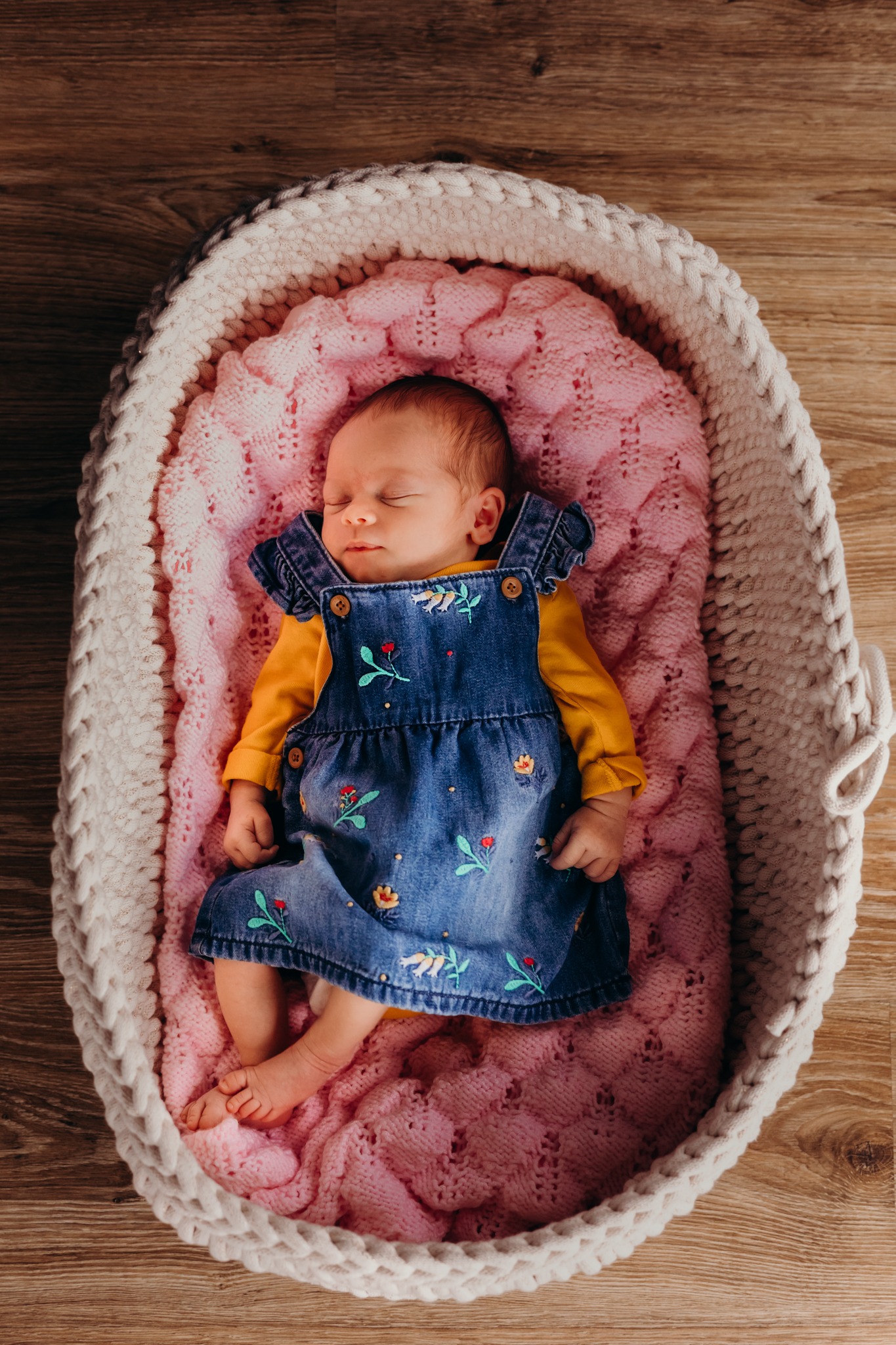 Noworodek śpiący w plecionym koszyku na różowym kocyku, ubrany w dżinsową sukienkę z haftowanymi kwiatami i żółtą bluzkę.