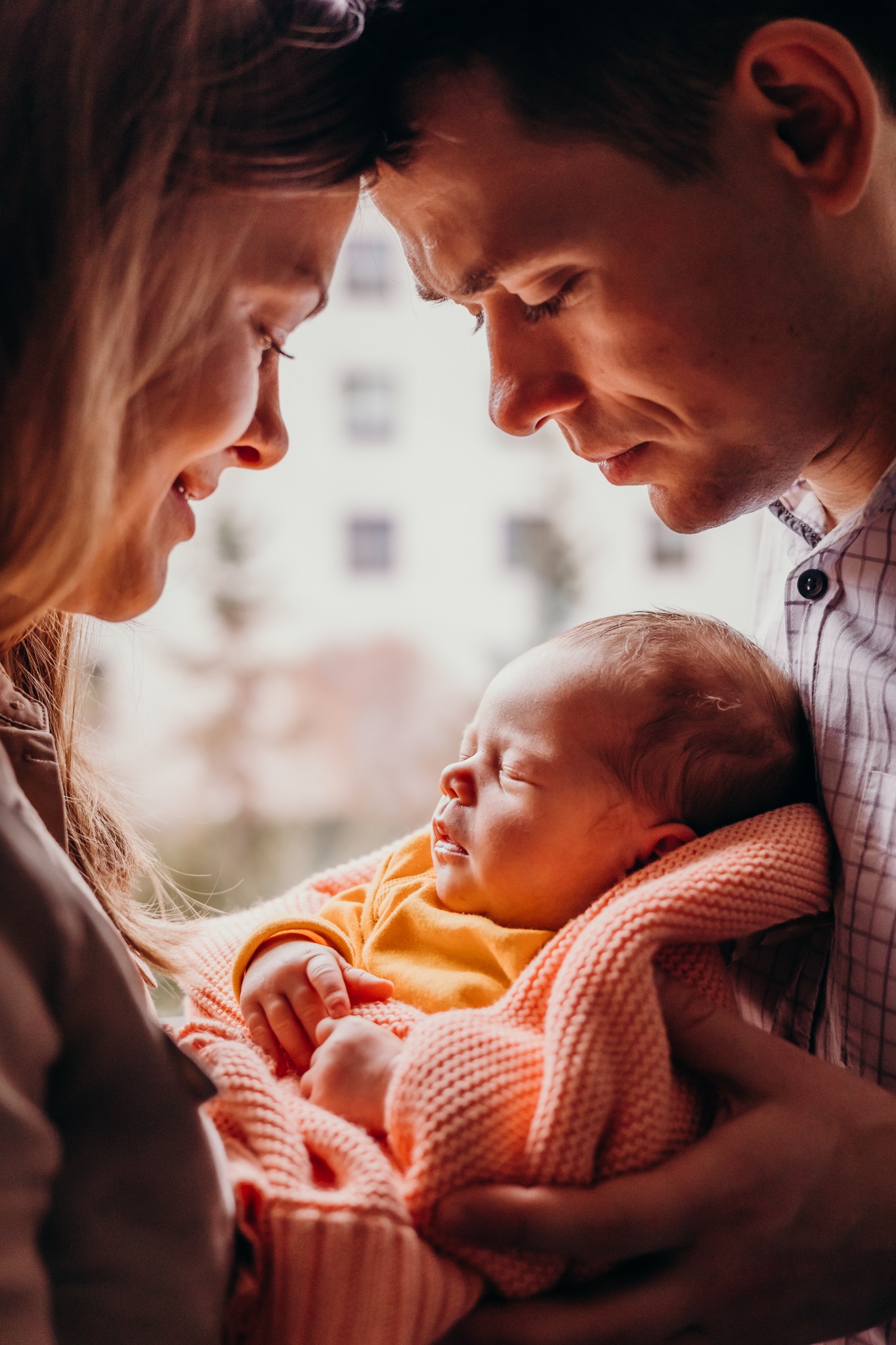 Rodzice patrzą z czułością na śpiące nowo narodzone dziecko owinięte w kocyk w odcieniach brzoskwini, delikatne światło okienne w tle.