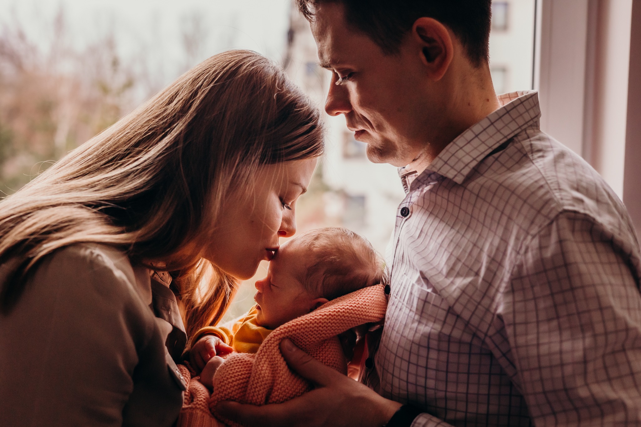 Rodzice czule tulą śpiącego noworodka przy oknie, matka delikatnie całuje dziecko w czoło, ciepłe światło podkreśla intymną atmosferę