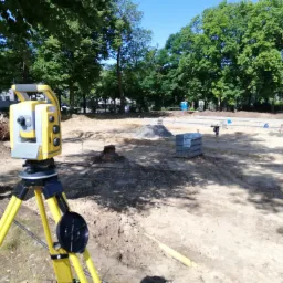 Żółty teodolit ustawiony na placu budowy, w tle widoczna praca ziemne, sterty materiałów i pracownik z taczką.