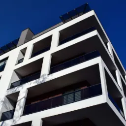 Nowoczesny budynek mieszkalny z białymi elewacjami i balkonami z ciemnymi balustradami, widziany z dołu na tle intensywnie niebieskiego nieba.